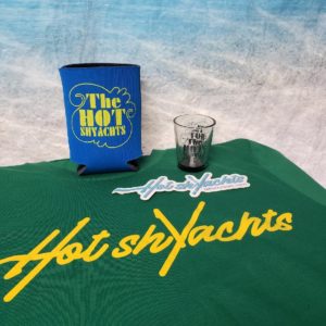 Hot ShYachts t-shirt, koozie, shotglass, and sticker displayed in photo against beach backdrop